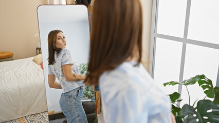 A young woman indoors posing playfully in front of a mirror set in a modern bedroom with natural light.