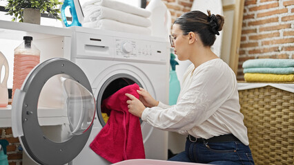 Young beautiful hispanic woman putting clothes in washing machine at laundry room