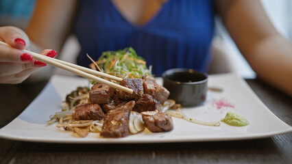 Young woman joyfully digs into her delicious gourmet beef meal, heartily using her chopsticks at a modern asian restaurant. she's enjoying her fresh japanese cuisine for an amazing lunch or dinner