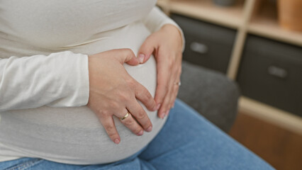 Young hispanic pregnant woman creating heart shape with hands on her belly, indoors at home.