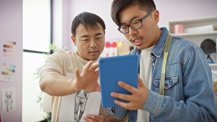Two asian men collaborating in a tailor shop with one holding a tablet, both focused on design details.