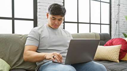 A young asian man focused on his laptop in a modernly decorated apartment living room.