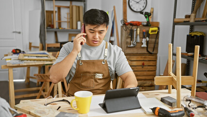 Fototapeta premium Chinese man in apron talks on phone at carpentry workshop with tools and tablet