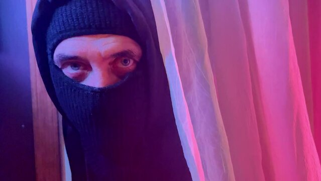 A robber burglar is breaking into a home with a black mask and weapon with red light shining on the curtain in the night darkness for a home surveillance or security concept. 