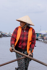 Vertical photo of a Vietnamese woman wearing a conical hat, she is navigating through the floating market in the mekong delta