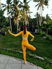 Woman in vibrant yellow outfit gracefully strikes yoga pose amid lush palm trees on tropical...