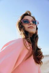 Stylish woman in pink coat and sunglasses gazing at the sun on the beach