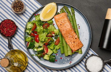 Healthy lunch with grilled salmon with asparagus and fresh tomato and cucumber salad, a row of lemon slice.