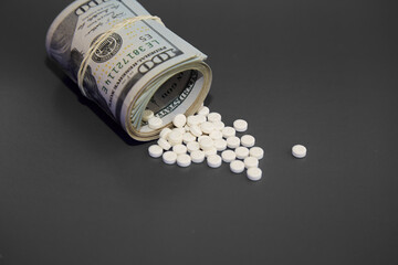 A rolled up wad of hundred dollar bills with white pills spilling out on a gray background, money for medicine, paid medicine, health insurance
