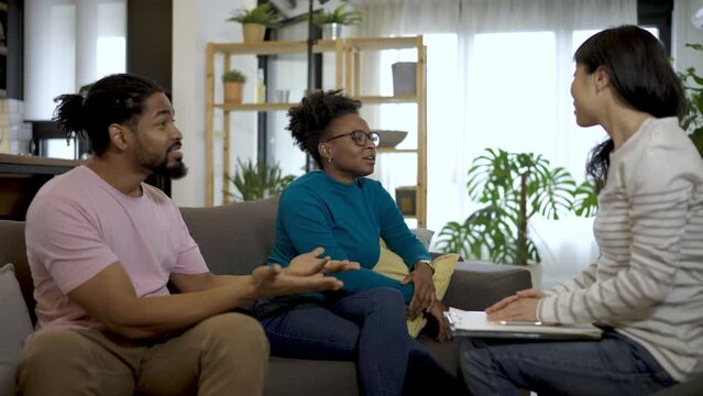 Young Black couple is seated in a cozy office, talking with an Asian female marriage therapist. The therapist listens attentively, offering guidance and fostering constructive communication between th