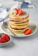 A stack of pancakes with strawberries and a chamomile flower, drizzled with icing on a plate, on a light background.