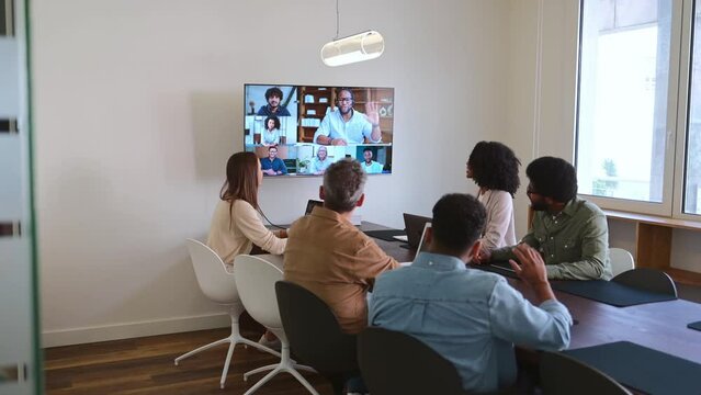 A group of diverse colleagues is gathered in a modern conference room, actively participating in a video conference call displayed on a large screen. Meeting showcases a collaborative work environment