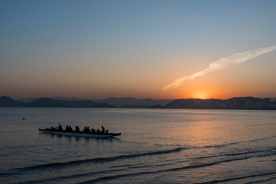 Silhouette of athletes leaving the beach paddling in a Hawaiian canoe during the sunset. Santos city, Brazil.