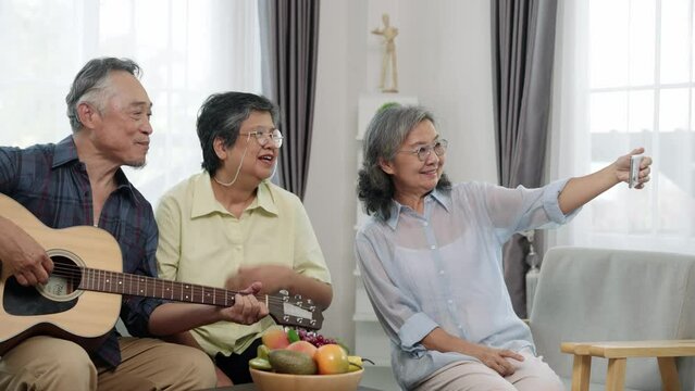 Handheld shot of happy elderly friends in a living room. One plays the guitar while another snaps a selfie, preserving their cherished moments of music and companionship.
