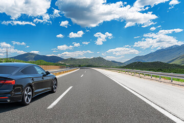 A black car drives along the highway against the backdrop of rocky mountains on a sunny day.