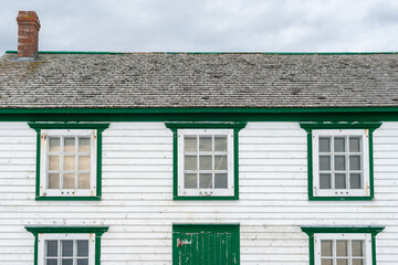 The exterior wall of a vintage white wooden building with green trim, grey cedar roofing shingles and solid wooden shutter door. The building is weathered and worn with a red brick chimney.