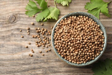 Dried coriander seeds in bowl and green leaves on wooden table, flat lay. Space for text
