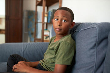 Worried black teenager in casual clothes sitting on couch in his room as punishment for bad...