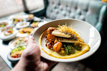 Close-up of a hand holding a plate with a gourmet dish featuring fish, asparagus, and garnish on a puree