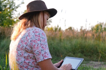 Back view of woman in hat using tablet in outdoor nature setting. Woman using tablet outdoors in nature during summer, back view. Casual summer activities with technology