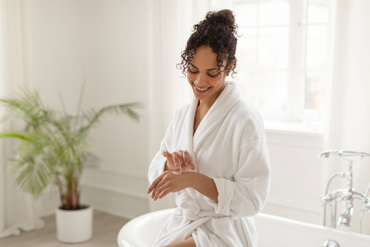 Young Black lady applying hand cream while sitting on bathtub in light bathroom interior, woman enjoy making beauty treatments