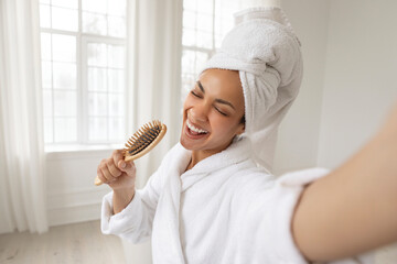 Happy black woman singing song holding hairbrush as microphone, having joy at free time in the morning. Young lady with towel on head after shower