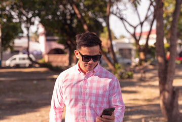 Youthful looking Asian man in his mid 40s with a neck tattoo, wearing sunglasses and a pink plaid shirt, and using his smartphone in an outdoor park setting.