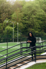 Smiling woman dressed in black activewear sitting on grass, engaging with her smartphone in a lush green park.