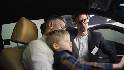 A confident middle-aged man with gray hair and a white shirt communicates with a car dealership assistant in a business suit while sitting with his little son in the interior of a modern car