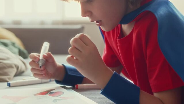 Chest up of Caucasian boy wearing red and blue superhero costume drawing on coloring page with crayons while playing at home at daytime
