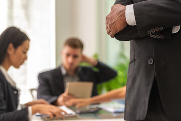 A black man in a suit stands with his arms crossed in front of a group of people.