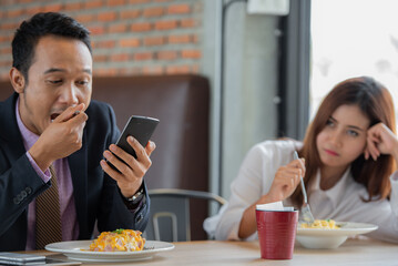 A man is looking at his phone while a woman is sitting across from him, looking bored.