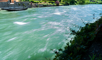 long exposure of the turquoise waters of a flowing river, flanked by verdant flowering banks; on the water stands a traditional river boat
