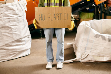 Young volunteer in gloves and safety vest holds sign that says no plastic while sorting waste.