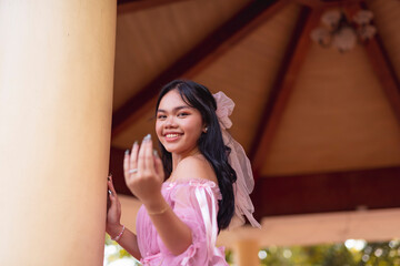 Fototapeta premium A young Asian woman in a pink off-shoulder dress at an outdoor gazebo in the park, inviting with a smile.