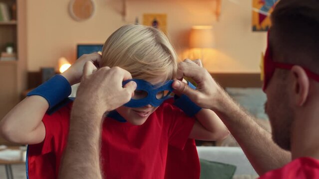 Over shoulder of dad helping tween son to wear blue superhero eye mask while having fun together in apartment