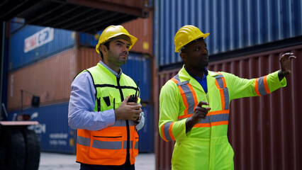 Two workers wearing hard hats and safety vests discuss logistics and operations at a shipping container yard with stacked containers in the background.  