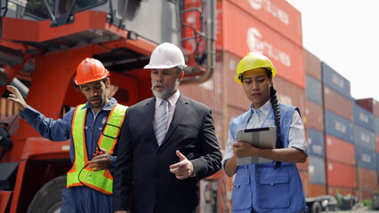 A manager in a suit and hard hat talks with two workers in safety gear at a busy shipping yard with containers in the background.