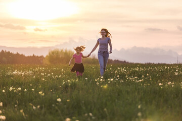 Happy and smiling mother and her little daughter running holding hands across a meadow in the beautiful setting sun.