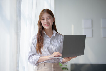 Smiling woman standing by a window, holding a laptop. Bright, airy environment suitable for work or study at home.