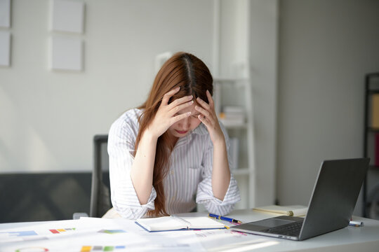 Stressed woman at work holding her head, overwhelmed with tasks, surrounded by documents and a laptop in a modern office.