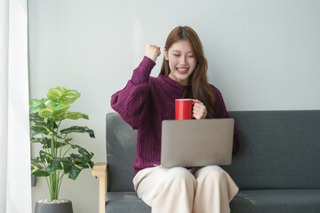 Woman celebrating success while holding a red coffee mug and using a laptop on a couch. Bright and...