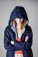A cheerful young woman in a blue hooded jacket over a sporty gray hoodie and bright orange top,...