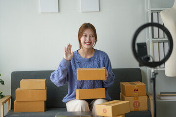Woman recording an unboxing video at home with a camera, smiling and waving, surrounded by multiple...