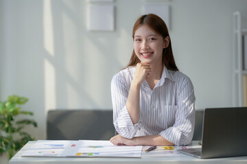 Young professional woman smiling at desk with documents and laptop in modern office, sunlight streaming through the window.