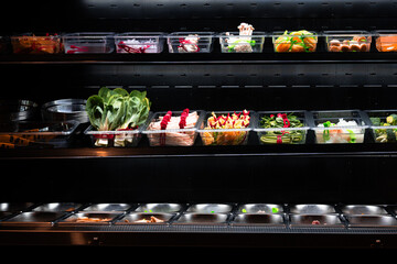 A colorful display of fresh vegetables and ingredients in clear plastic containers at a market or restaurant. The neatly organized setup features bok choy, mushrooms, bell peppers, and greens, showcas