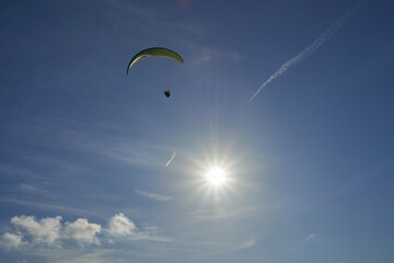 A paraglider soars in a clear blue sky, the sun creating a lens flare. The canopy is green and...