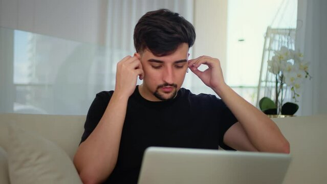 Young Man Using Laptop with Earbuds at Home. Guy with dark hair sits on a couch, wearing earbuds and using a laptop, remote work. Student studying online through laptop at home