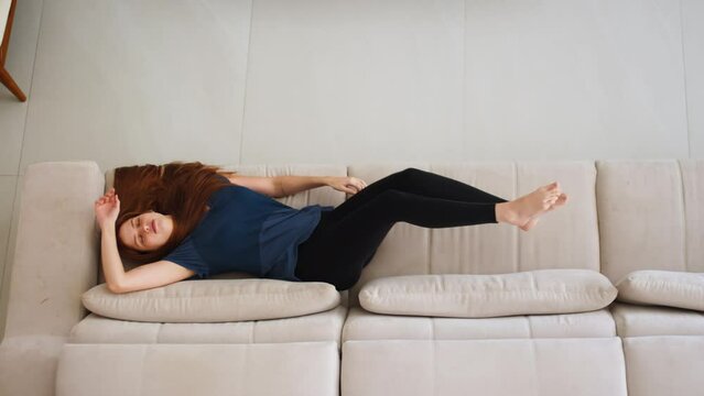 Young Woman Relaxing on Couch with Eyes Closed. Girl with red hair lies down on a beige couch, closes her eyes to relax and enjoy her free time. Leisure and relaxation at home