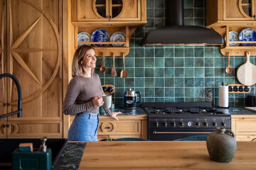 A woman in a beautiful kitchen surrounded by wooden furniture walks with coffee and thinks